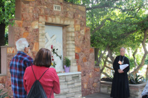 A Passionist brother stands beside an outdoor stone shrine marking the Eighth Station of the Cross, speaking to two people facing the shrine adorned with flowers.