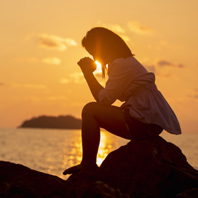 Silhouette of a woman kneeling in prayer on rocky ground by the water’s edge, with the sun setting over the horizon in the background.