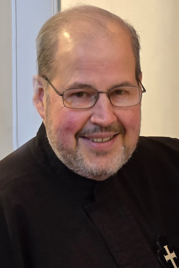 Smiling Passionist brother wearing glasses and a black habit with the Passionist emblem, photographed indoors.