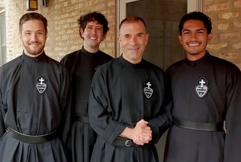 Four smiling Passionist priests wearing black habits with with the Passionist sign