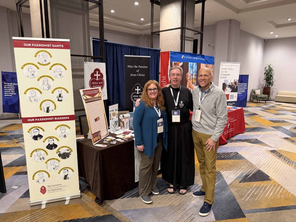 Three smiling people in front of a trade show booth for the Passionists