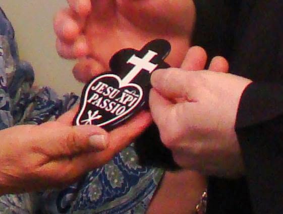 Hands present a heart-shaped Passionist Sign featuring a white cross, the words "Jesu XPI Passio," and three nails, symbolizing the Passion of Jesus Christ and the mission of the Passionists.