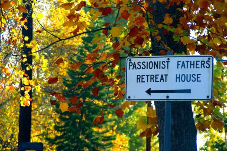 A directional sign hanging on a tree amidst fall foliage points to a Passionist retreat house