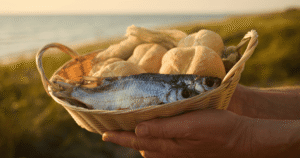 Hands in front of a body of water offering a basket of loaves of bread and fishes