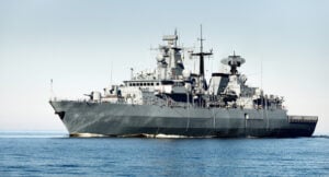 A large gray naval warship sails across calm blue waters, with radar equipment and antennas visible on its deck.