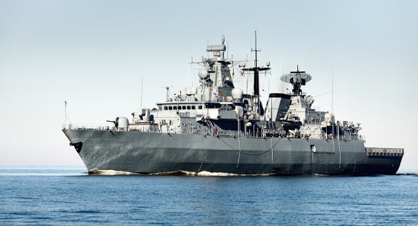 A large gray naval warship sails across calm blue waters, with radar equipment and antennas visible on its deck.