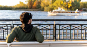 A young man sits on a waterfront bench, looking out across a river as a boat passes in the distance.