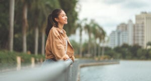 A young woman smiles while leaning against a railing beside a river, with palm trees and city buildings in the background.