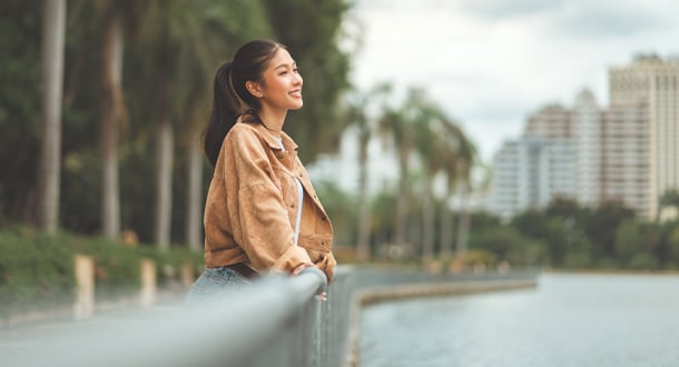 A young woman smiles while leaning against a railing beside a river, with palm trees and city buildings in the background.
